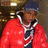 A person in a red jacket with white trim and dark blue cap stands against a blurred background of warm-toned lights.