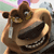 Pixelated cartoon animal with brown fur, large black nose, and wide eyes, holding a white object near its face against a blurred background of pastel colors.