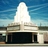 Static pixelated image of a white building with a tall central tower under a blue sky, featuring a dark rectangular entrance and horizontal architectural lines.