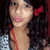 A person with long dark curly hair and a red bow in their hair, wearing a gray patterned top with red straps.