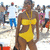 A woman in a yellow one-piece swimsuit stands on a beach with a crowd in the background.