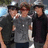 Three figures stand side-by-side in a pixelated image; the central figure wears a gray shirt and dark pants, flanked by two others in darker clothing against an indistinct background.