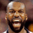 A pixelated close-up of a person with a bald head, dark beard, and open mouth showing teeth, wearing a dark jersey with white and blue trim against a blurred background.