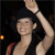 Pixelated figure with dark hair and light skin raises one arm against a dark background, wearing a dark sleeveless top with a red strap visible on the left shoulder.