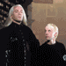Two figures in black attire stand side-by-side against a textured backdrop, with green text overlaying the scene.