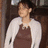 Pixelated image of a person wearing white long-sleeve shirt and brown apron, standing against dark background.