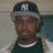 A person wearing a dark cap with white 'NY' logo, dark green shirt, and light-colored jacket with patterned sleeves, against a two-tone wall background.