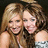 Two women with long brown hair in off-shoulder silver-patterned tops pose closely together against a dark background.