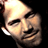 Pixelated static image of a smiling man's face with light skin tone, dark hair, and visible facial features against black background.