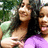 Two people embrace outdoors; one wears a black top and purple garment, the other a light green top, with blurred green foliage in the background.