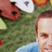 Pixelated image of a person with light skin and brown hair wearing a green top, with a red object and blue background visible.