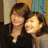 Two people with brown hair pose closely together, one wearing a dark top and the other in light-colored clothing against a blurred background.
