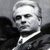 Static grayscale pixelated image of a person with short light hair, dark eyes, and a neutral expression wearing a dark jacket over a white shirt against a gray background.