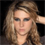A static pixelated image of a person with light brown wavy hair, dark eyes, and a neutral expression against a dark background.