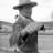 Pixelated grayscale image of a figure wearing a wide-brimmed hat and dark clothing, standing against a light background with indistinct shapes.