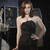 A woman in a strapless black dress poses with one hand on her hip against a dark background with blurred lights.