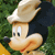 Pixelated cartoon character with large ears, light brown face, and white hat, set against green foliage background.