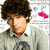 A person with curly brown hair wears a light gray sweater over a white collared shirt, standing against a patterned background with pink shapes.