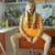 A person with long light hair sits on a red chair holding an orange object, positioned against a blurred background.