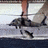 Pixelated static image of a dark-colored aircraft in flight over water with a light sky background.