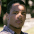 Static 50x50 pixel icon showing a close-up of a person's face with dark hair and light skin, wearing a blue shirt against an outdoor background with green foliage and white elements.