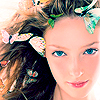 A close-up of a person with long brown hair adorned with colorful butterfly-shaped hair clips, blue eyes, and a nose ring, set against a bright background.