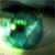 A pixelated green circular icon with a dark center and light green highlights, set against a blurred background with green and brown tones.