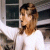 Pixelated image of a person with light brown hair wearing a white shirt, standing in front of dark rectangular panels and a blurred background.