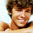 Pixelated close-up of a person with curly brown hair, light skin tone, and visible teeth in a smile against a pale blue background.