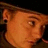 Pixelated close-up of a person's face with light brown hair, visible ear, and dark background, rendered in warm orange-brown tones.