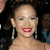 A pixelated close-up of a person with dark hair, wearing large dangling earrings and red lipstick, against a dark background with blurred figures.