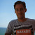 Pixelated static image of a person wearing a gray shirt with dark horizontal stripes and a red rectangular patch, standing against a light blue sky and water background.