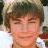 A pixelated image of a person with light brown hair and closed eyes, wearing a white shirt with red accents, featuring a bright pink heart symbol on the right side.