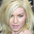 Pixelated close-up of a person with light-colored hair, visible eyes, nose, mouth, and part of a shoulder against a blurred background.