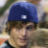 Pixelated static icon showing a person wearing a blue cap with a white square detail, partially visible face, and blurred background elements.