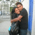 Two people embrace in a close hug, one with short dark hair and the other with light brown hair, against an outdoor background with blue vertical structures.