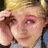 Pixelated close-up of a person with light hair holding their hand near the face, wearing pink eyeshadow and a dark top.