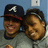 Two pixelated figures with brown hair and light skin, one wearing a dark cap with a white 'A' symbol, both smiling against a blue background with a white square element.