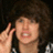 Pixelated close-up of a person with dark curly hair holding a microphone near their mouth, wearing a checkered shirt.