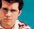 Pixelated close-up of a person with short dark hair against a solid teal background, wearing light-colored clothing.