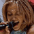 Static 50x50 pixel icon showing a close-up of a person with light brown hair wearing a dark top, partially visible blue garment, and red lip color against a blurred background.