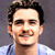 Static pixelated portrait of a person with dark hair and light skin wearing a blue shirt against a gray background.