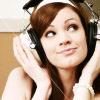 A person with brown hair wearing black headphones and a white headband, holding the headphones with both hands against a light brown background.