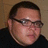 Pixelated close-up of a person with short dark hair, wearing glasses and a dark shirt against a neutral background.
