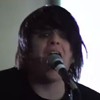 A person with dark hair sings into a microphone, wearing a dark shirt against a light background.