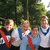 Group of people in white and blue shirts with red sashes standing outdoors under trees, some faces blurred.
