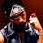 Pixelated static image of a person with dark hair wearing a black shirt and light-colored jacket, holding an object in their right hand against a brown background.