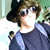 A person with brown hair and sunglasses wears a dark shirt, blue backpack strap visible, against green foliage background.