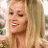 A pixelated close-up of a person with long light brown hair, wearing red lipstick, and smiling with visible teeth against a blurred background.