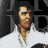 Pixelated static icon of a stylized figure in brown tones with dark sunglasses against a gray and black background, appearing to hold an object near the face.